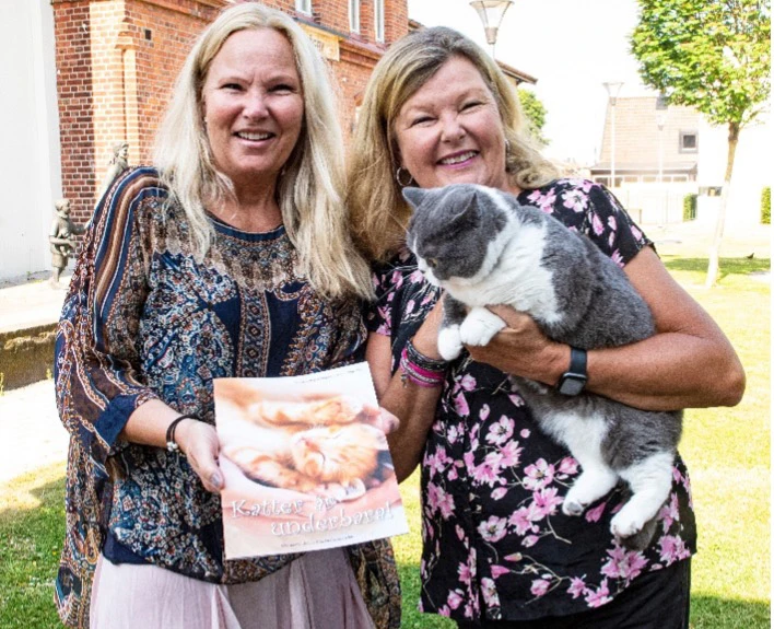 Eva Hellman Persson och Susanne Hellman Holmström står bredvid varandra och håller broschyren och en katt.