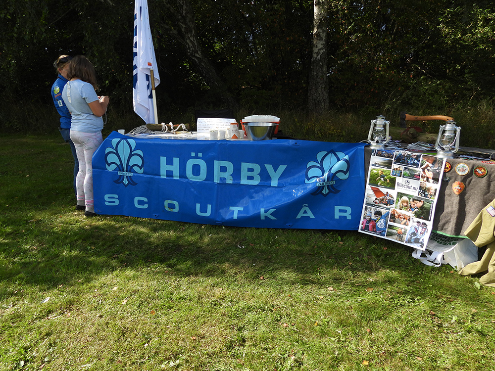 Bord utomhus med banderoll från Hörby scoutkår. Två personer står vid bordet men tittar inte in i kameran.